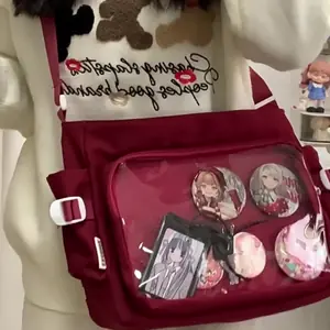 A person wearing a burgundy red Ita messenger bag with a transparent window displaying anime pins and charms, paired with a casual aesthetic outfit